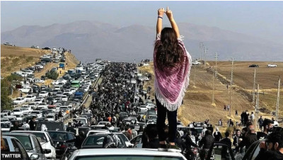 An Iranian woman, unveiled, stands on top of a car with her back to the camera and raising her arms to the crowd of people and vehicles on the highway behind her in protests about Mahsa Amini's death in police custody on 16 September 2022.